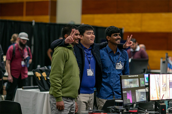 Three male students stand in front of a conference booth with computer screens displaying a research project.
