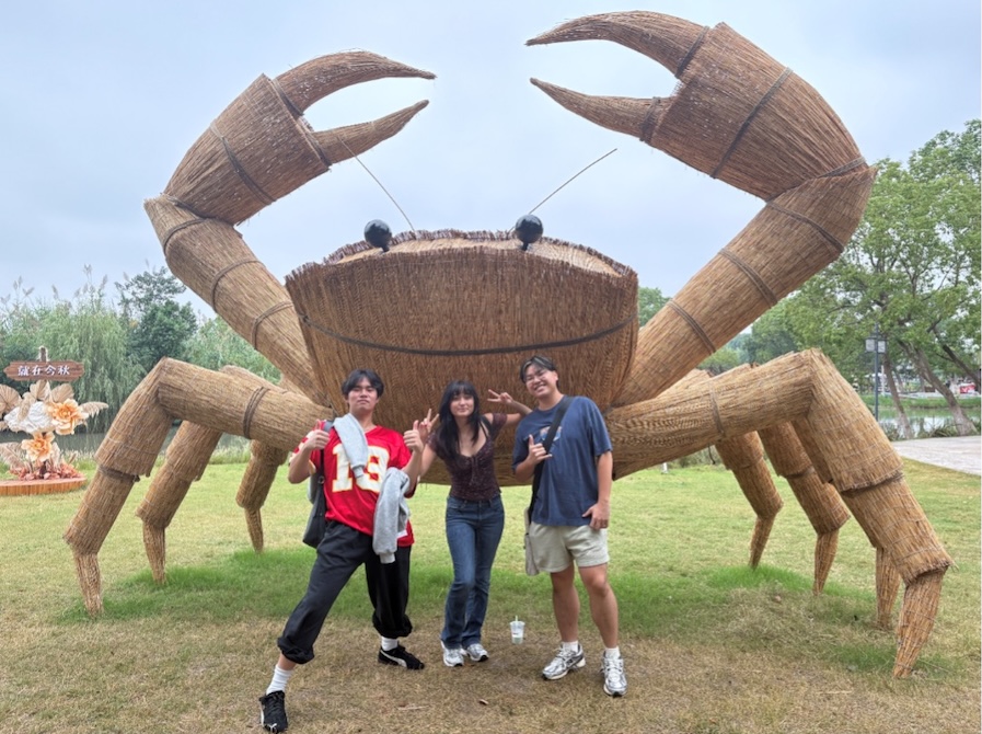 Three students stand in front of a 15 foot high crab sculpture made of wood. 