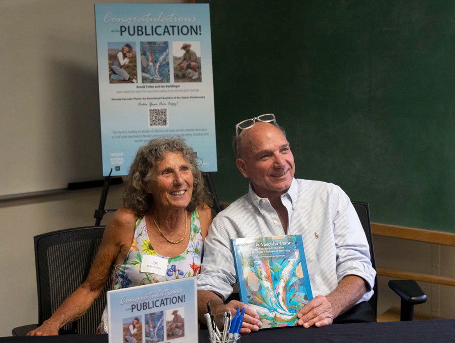 Two people sit behind a desk, one of whom is holding a book, smiling.
