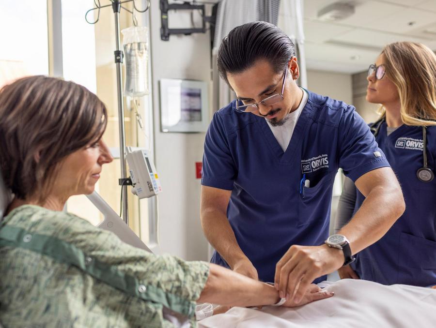Healthcare professional in Orvis School of Nursing scrubs checking on a patient in a hospital room, with another professional observing.