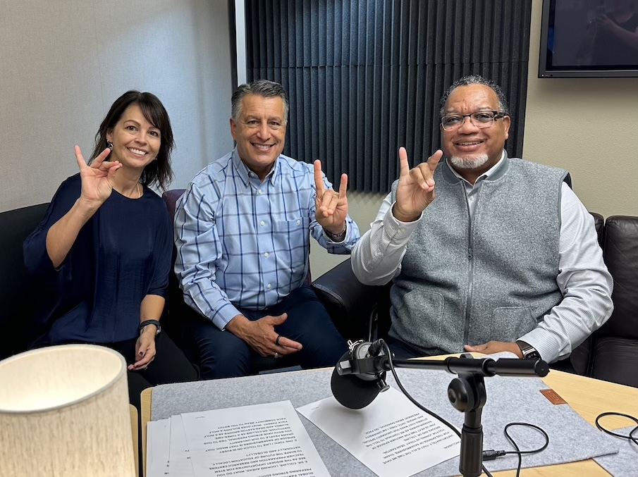 Mandi Collins sits next to President Sandoval and Dean Easton-Brooks in the podcast studio, all hold up Wolf Pack hand signs.