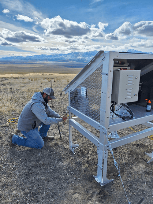 Person adjusting equipment at a weather monitoring station in an open field with mountainous background.