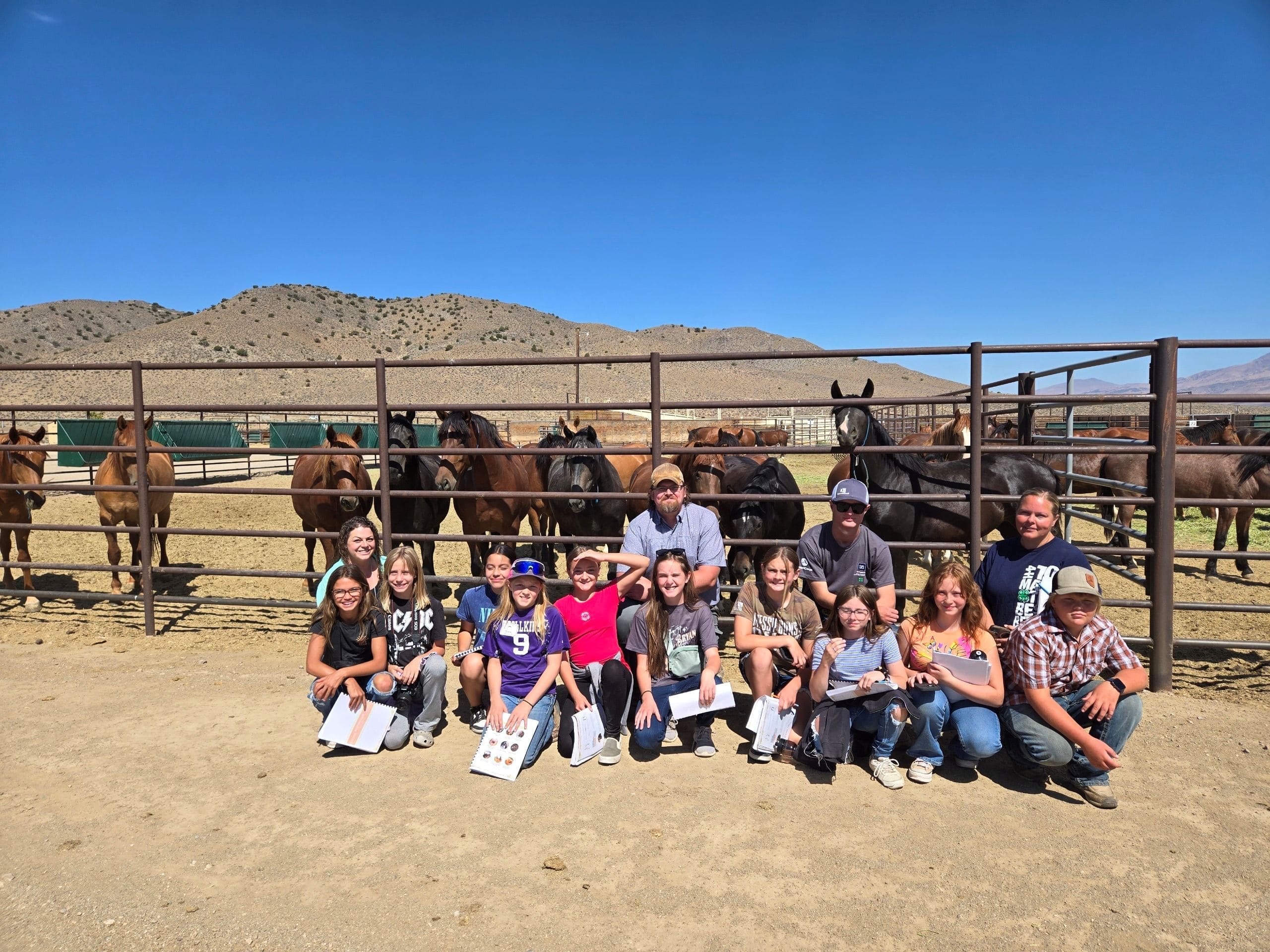Adults and children pose in front of a herd of mustangs across a bar fence.