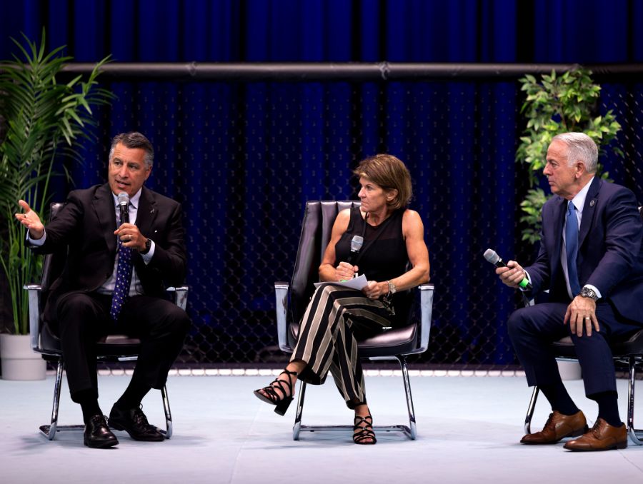 Nevada Governor Joe Lombardo, Staci Alonso, and University of Nevada, Reno President Brian Sandoval sit on stage during a panel discussion. Governor Lombardo gestures while speaking into a microphone, Alonso listens with notes in hand, and President Sandoval holds a microphone as he prepares to respond. The stage is set with potted plants and a blue curtain backdrop, creating a professional yet conversational atmosphere.