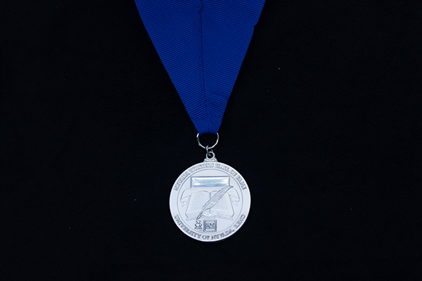 Silver medal attached to a blue ribbon featuring a ribbon, book, quill and ink well in the design. It has a Nevada block N and the words Nevada Writers Hall of Fame University of Nevada, Reno on one side.
