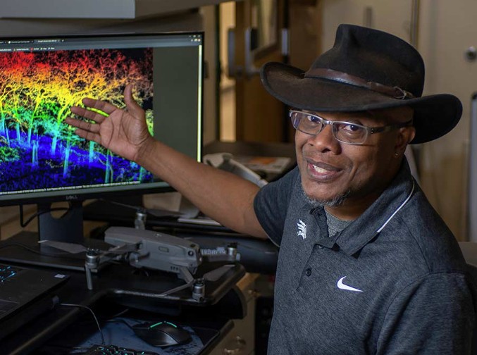 A man wearing a fedora hat points at a computer screen displaying illuminated objects while facing the camera.