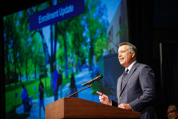 President Sandoval speaking at the podium with an image of Student Enrollment information on the PowerPoint behind him.