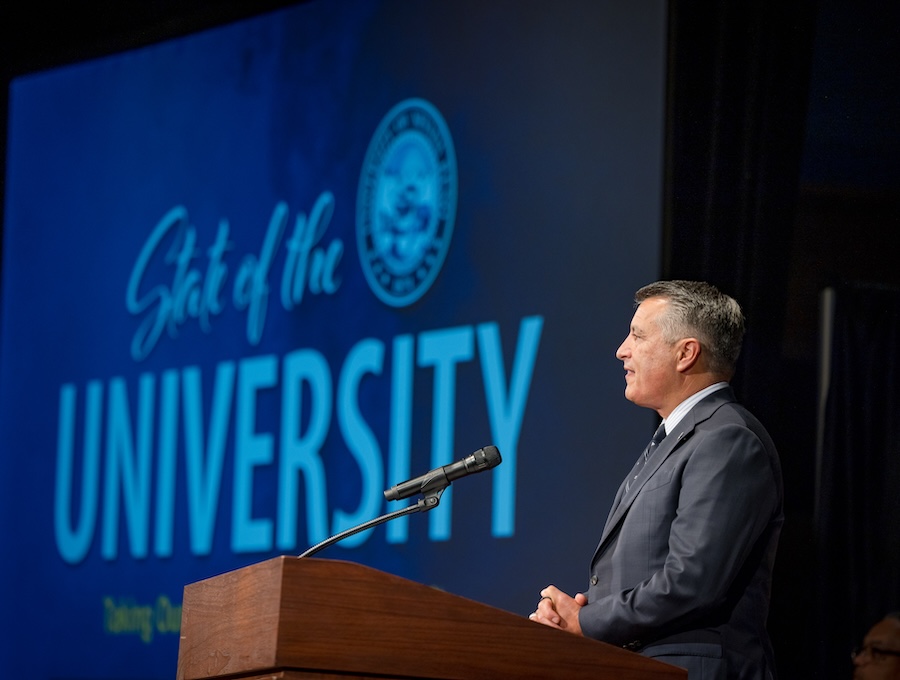 President Sandoval on stage during the address with the State of the University logo on a PowerPoint screen behind him.