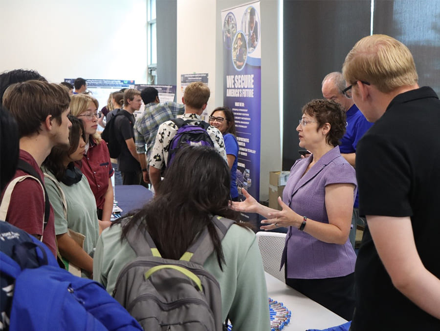 A woman at display table talks with a group of five students while other people in the background talk at other tables.
