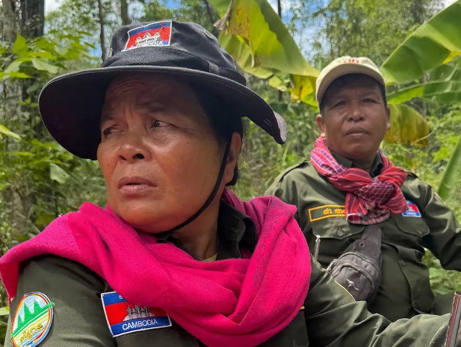 Two men dressed in fatigues with bright scarves around their necks sit in the forest.