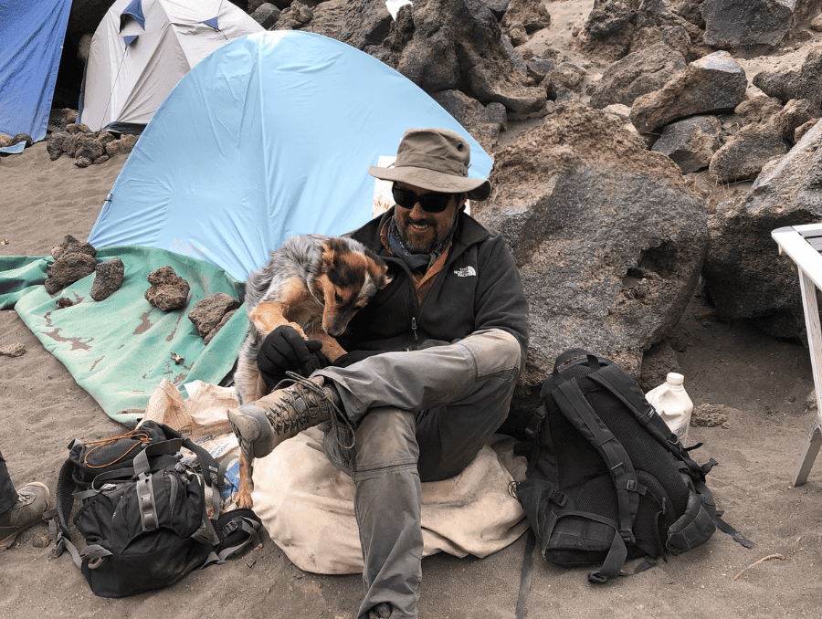 Chris Morgan sitting on a chair in front of a campsite, with a dog on his lap. 