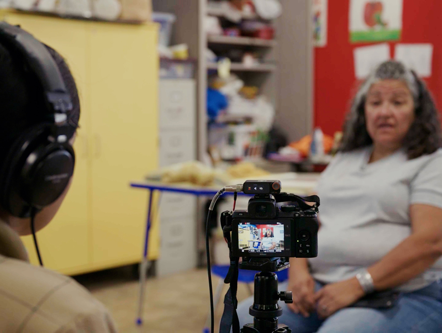 A person seated behind a camera facing another person who is being interviewed on camera for an article. 