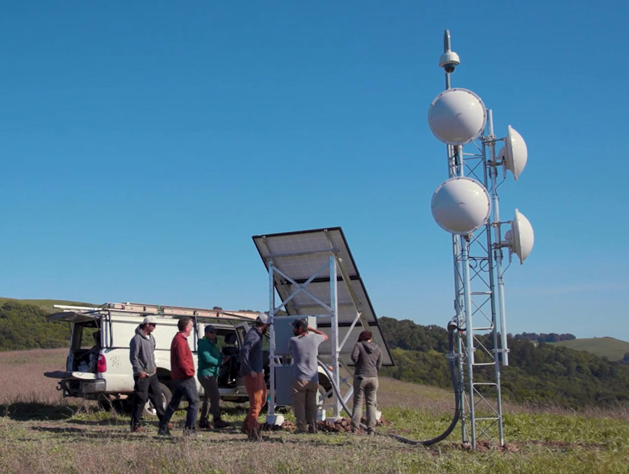Six people walking around a truck next to a large solar panel and satellite dishes.