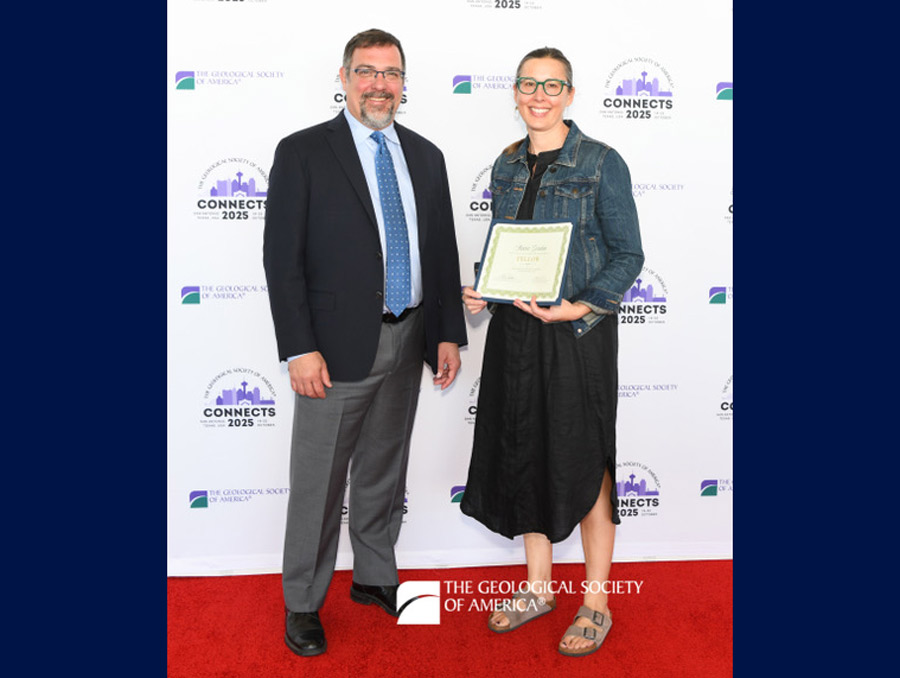Stacia Gordon stands on a rocky outcropping overlooking a valley surrounded by stunning, snow-capped mountains.