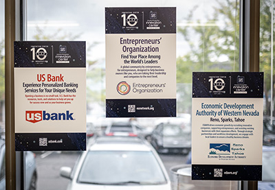 Three promotional posters displayed on a window, featuring logos and information for US Bank, Entrepreneurs' Organization, and Economic Development Authority of Western Nevada.