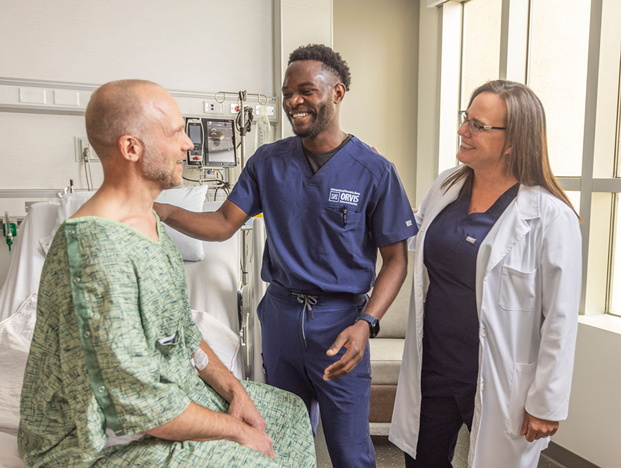 A patient in a hospital gown sits on a bed with a nursing student place their hand on their shoulder. A doctor of nursing in a lab coat stands smiling to the right.