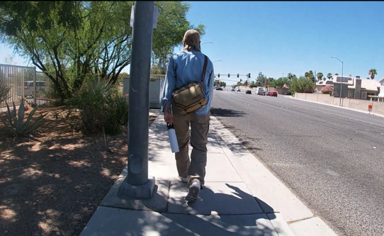 A person with a hat and a bag across their shoulder walking along on a street sidewalk on a sunny day.