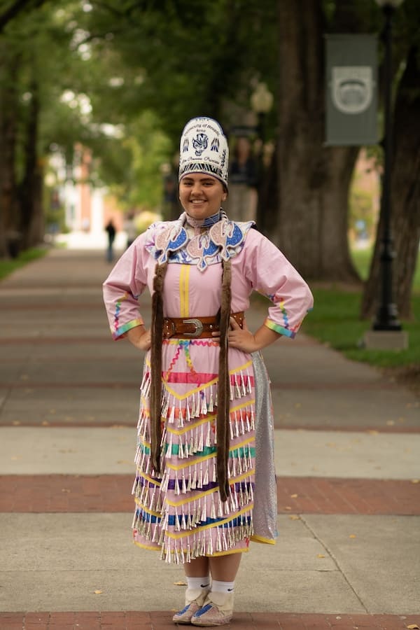 Riata standing on the University's quad wearing a traditional Native Powwwow dress.