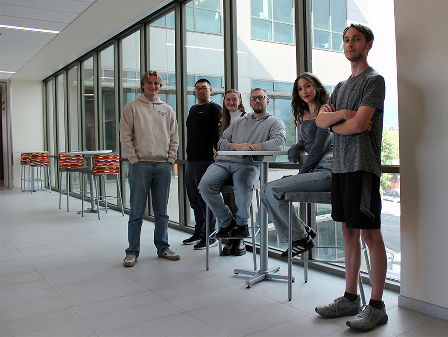 Five people in a hallway, one seated, a wall of windows in the background.