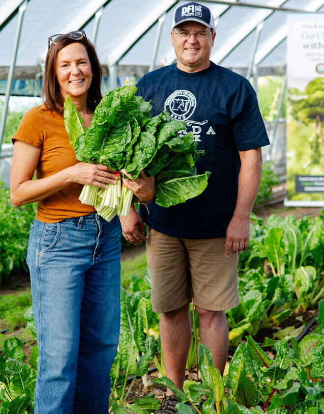 A smiling man stands beside a smiling woman who is holding a freshly harvested bunch of leafy vegetables.