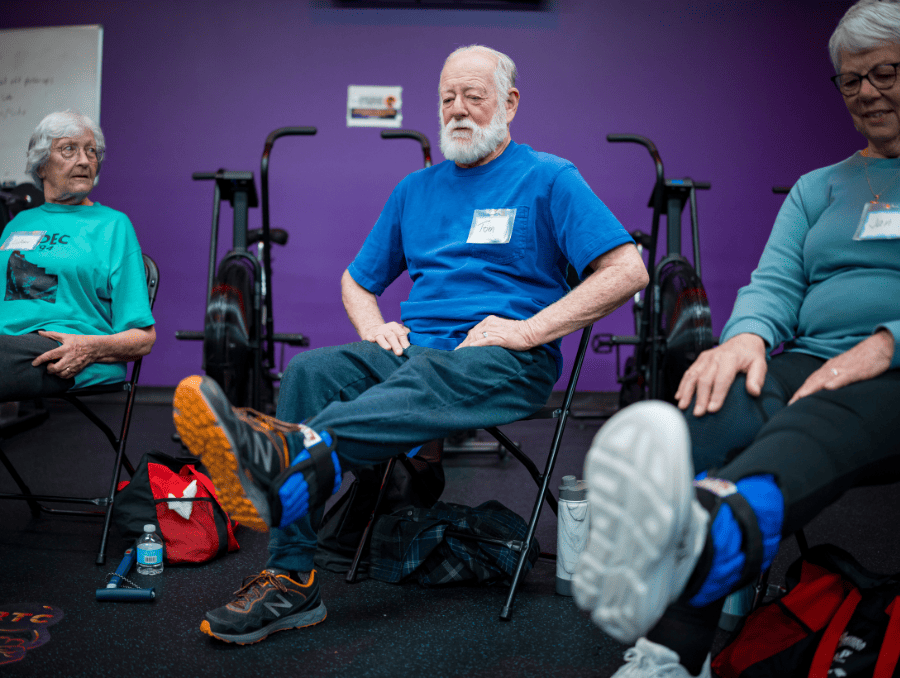 Three older adults stretching their legs while wearing weights inside a gym.
