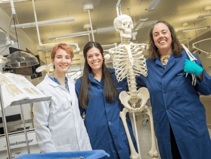 Three women standing with a skeleton in a lab.