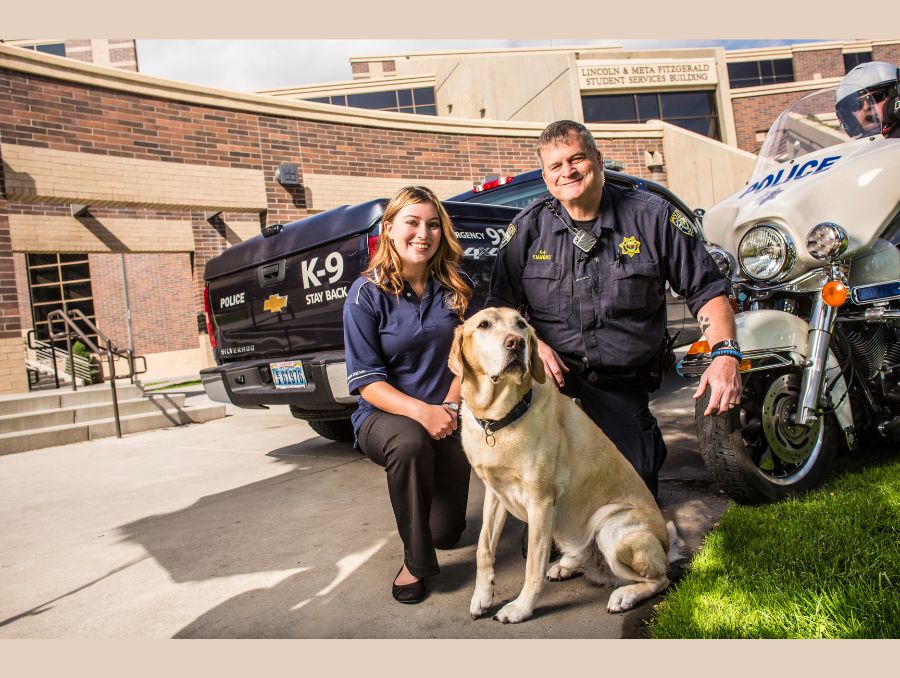 Two officers and a clerk with a dog posing in front of a cop car.