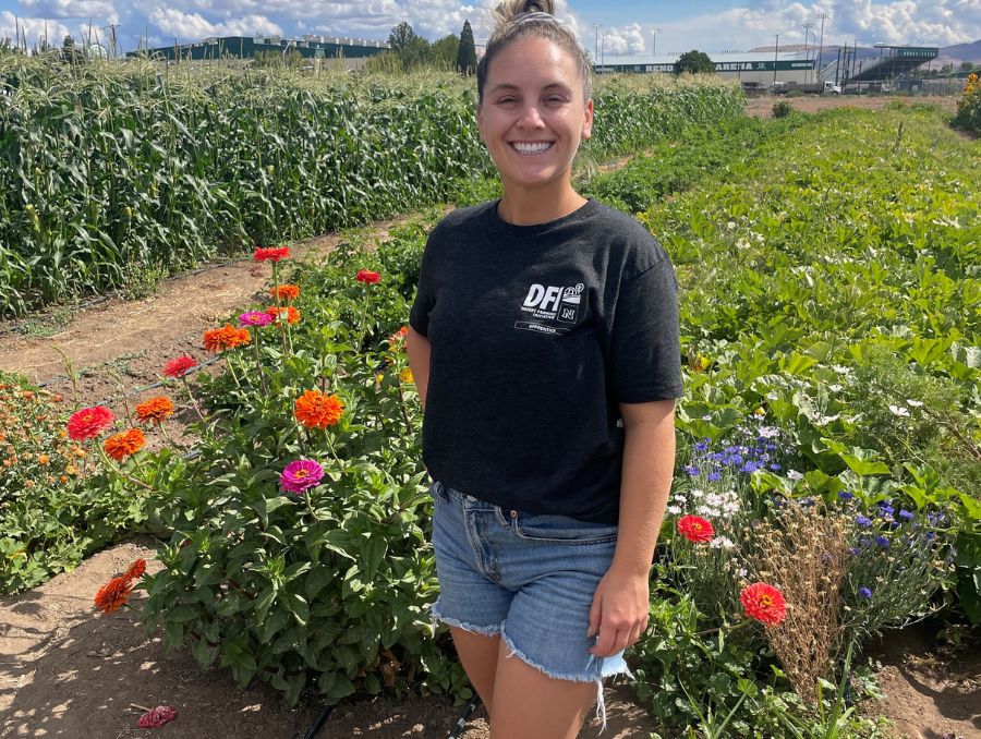 A smiling woman poses amid a farm with green plants and flowers.