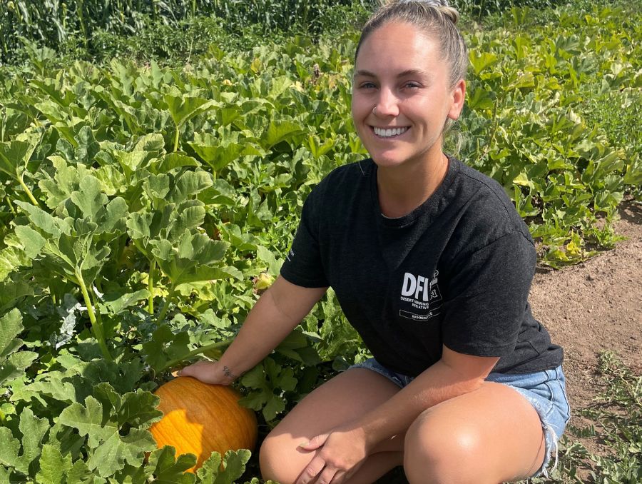 A smiling woman crouches in a pumpkin field holding an orange pumpkin.