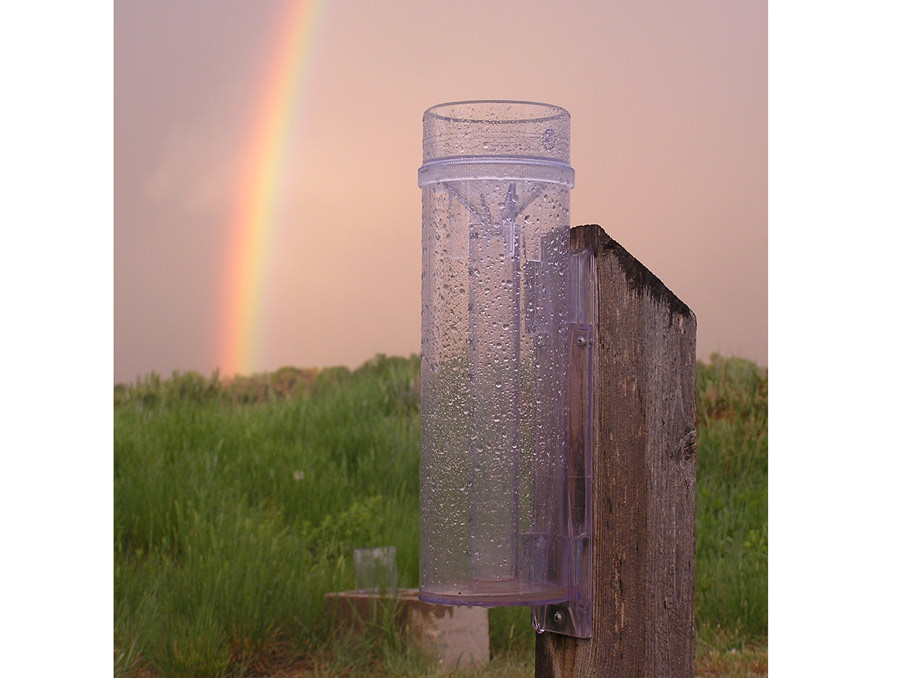 A rain gauge affixed to a wooden post with drops of water on it. Green foliage and a rainbow are in the background.