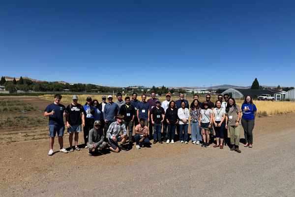 A large group of people stand outside on a bright sunny day in front of some fields, smiling for a photo.