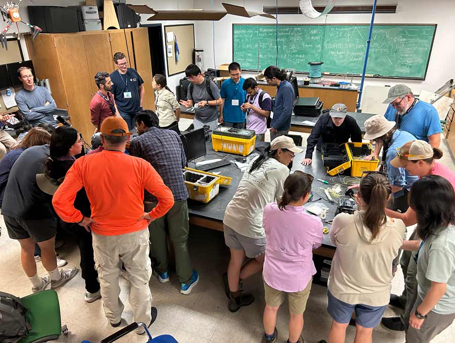 Several groups of people stand around desks in a classroom with yellow technology kits in front of them.