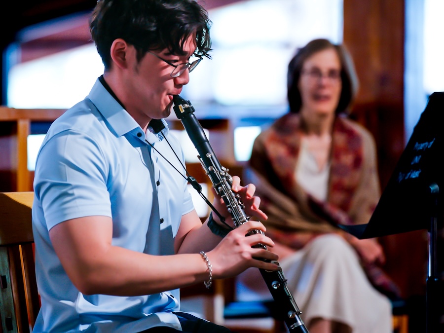 Student musician playing clarinet during a chamber music performance, with audience members watching in the background.