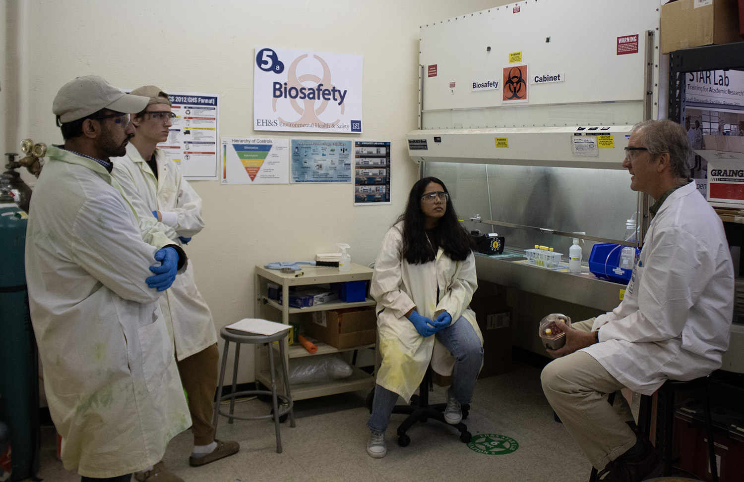 An instructor speaks to a group of three students about lab safety engineering in front the biosafety cabinet.  
