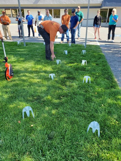 A man sticking white plastic cones on a grassy patch of land with a group of men and women watching.  