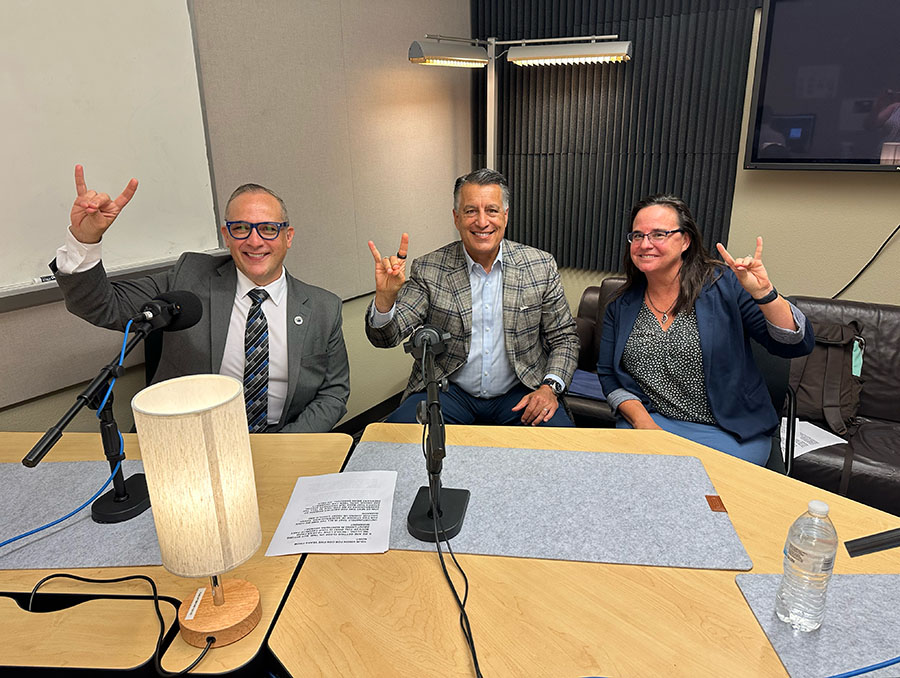 Brian Sandoval sitting next to Charlie Yingling and Shannon Richard in the podcasting studio holding up Wolf Pack hand signs.