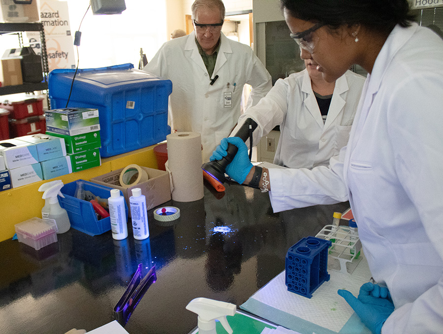 A student and two instructors look over a glowing spill using a black light. 