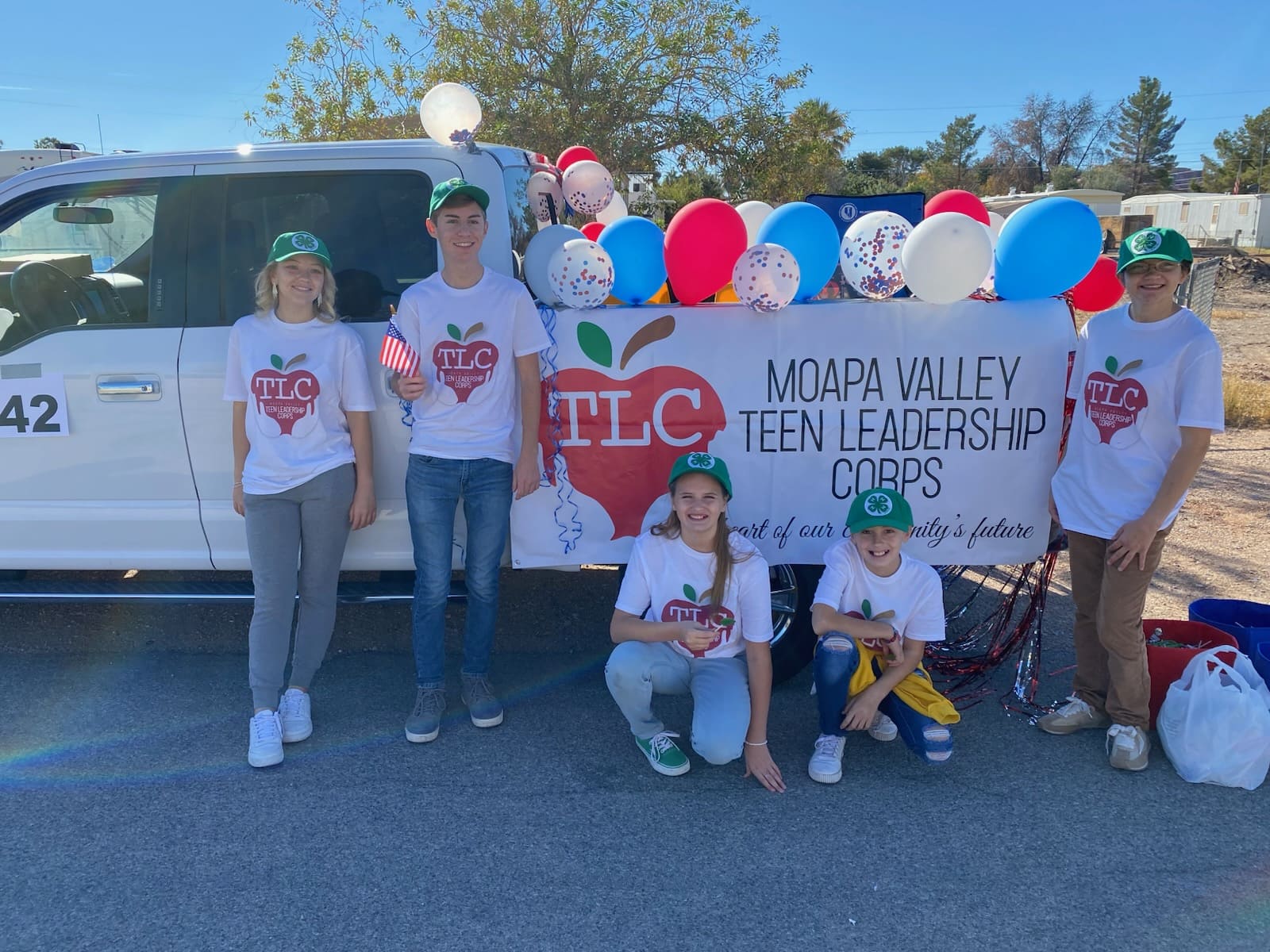 A group of boys and girls in white t-shirts and green caps posing in front of a white truck with colorful balloons in the back.
