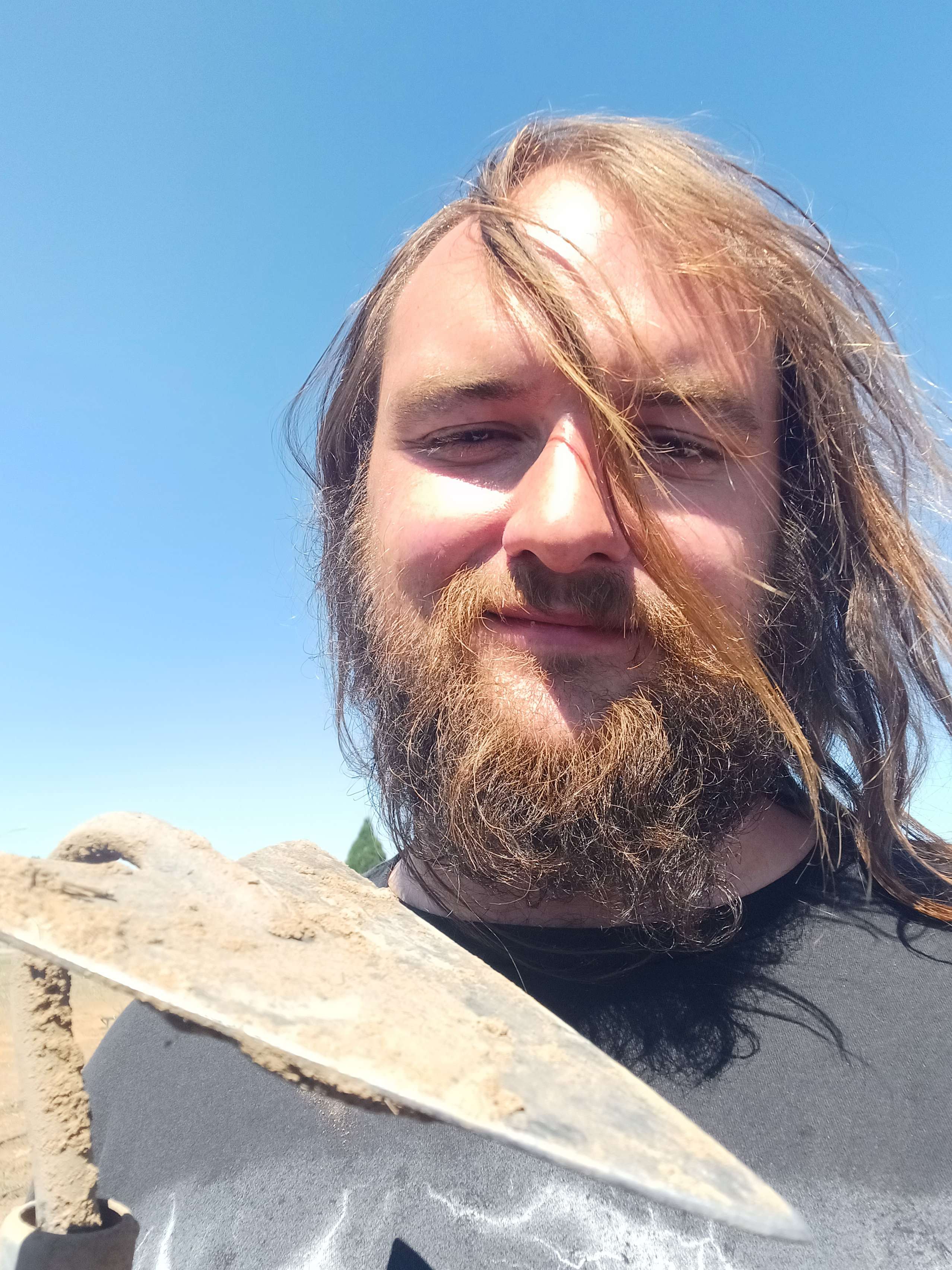 A long-haired man with a bushy moustache holding a digging hoe over his shoulder. 