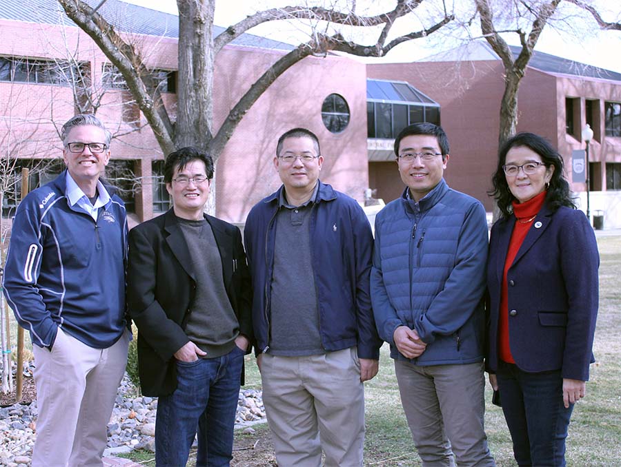 Four men and one woman stand in a line in front of a grassy area, trees and a red brick building.