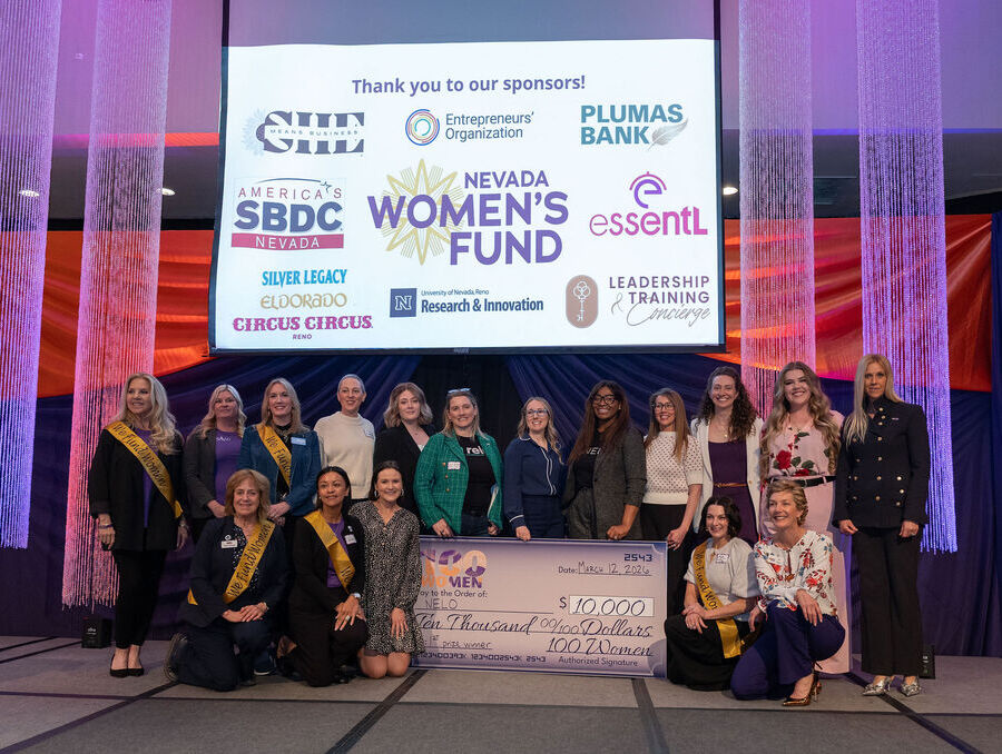 A group of 100 Women Pitch Competition participants pose on stage holding a $10,000 check, with event sponsors displayed on a large screen behind them.
