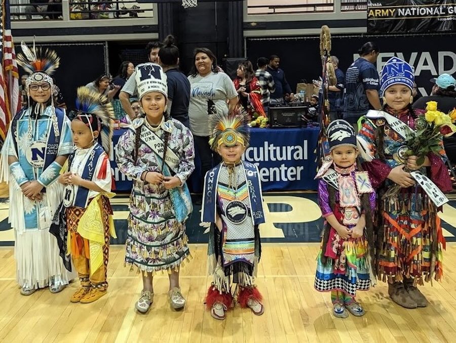 Children dressed in traditional Native American regalia pose on a basketball court.
