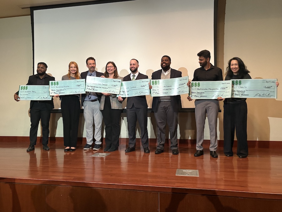 Winners of the competition stand on stage, holding large checks of their monetary winnings, with Graduate School dean Markus Kemmelmeier.