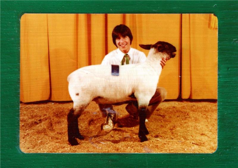 In a barn, a young, smiling, long-haired Brian Sandoval in 4-H dress crouches down to pose for a picture next to his blue-ribbon winning 4-H lamb.