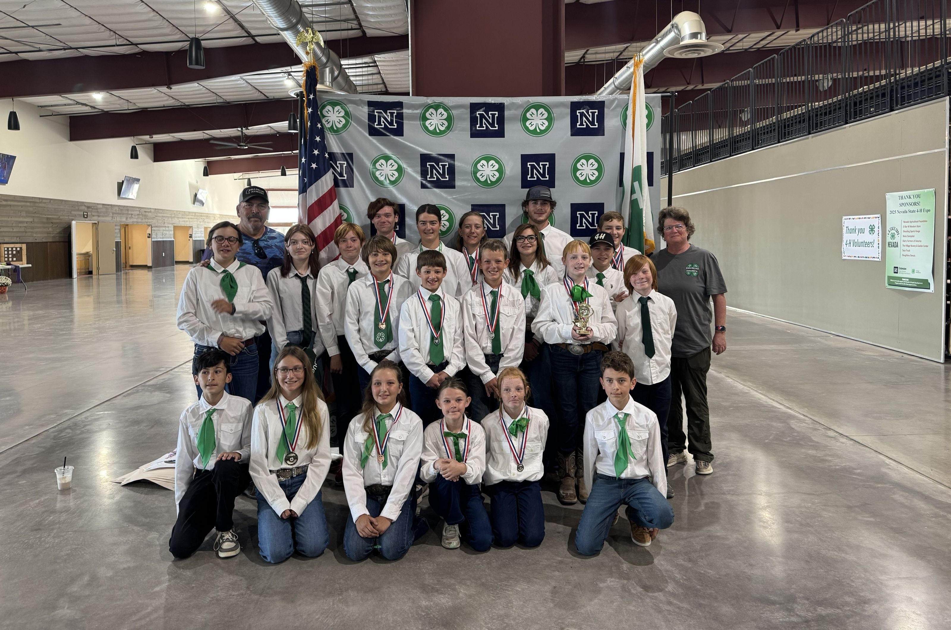 20 4-H youth and 2 4-H volunteers pose for a group photo in front of a University 4-H banner bookmarked by the U.S. and 4-H flags. The 4-H youth are wearing 4-H dress. Many are also adorned with medals. One is holding a trophy.