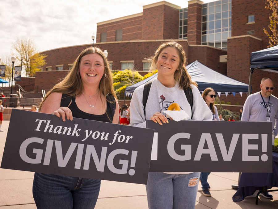 Two students stand smiling on the University of Nevada, Reno campus, holding “Thanks for Giving” and “I Gave” signs. 