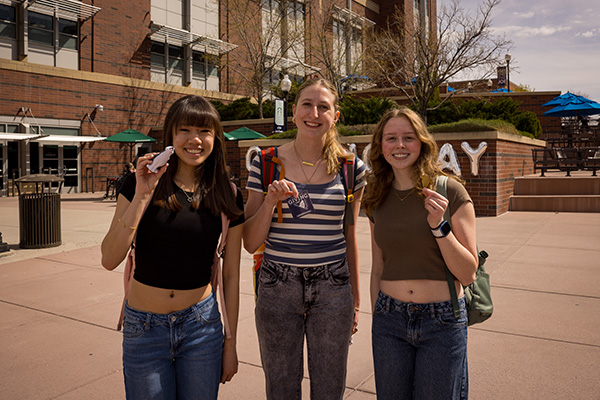 Three students display Giving Day stickers, Giving Day magnets and Giving Day &ldquo;stress wolves&rdquo; outside of the Joe Crowley Student Union. 