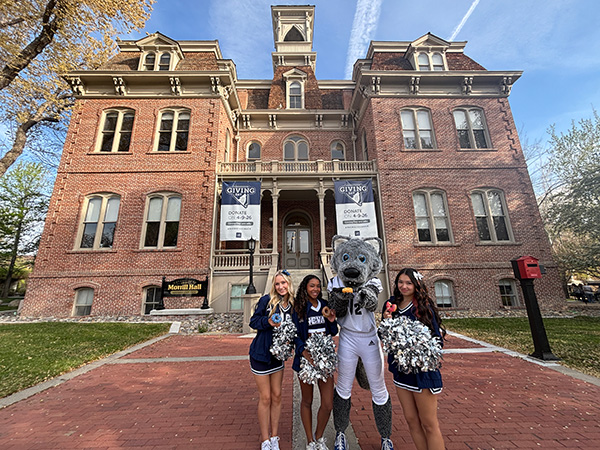 Three Nevada cheerleaders and Wolfie Jr. mascot stand in front of Morrill Hall Alumni Center framed between two Giving Day banners.  