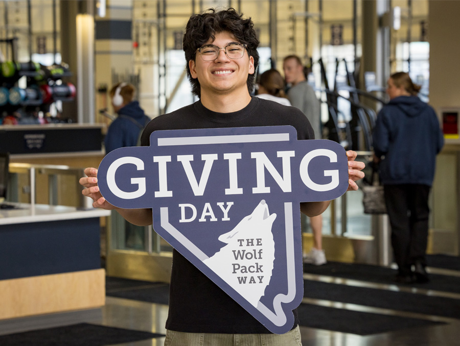 A student smiles and holds a sign with in the shape of Nevada with a Wolf on it that reads "Giving Day the Wolf Pack Way."