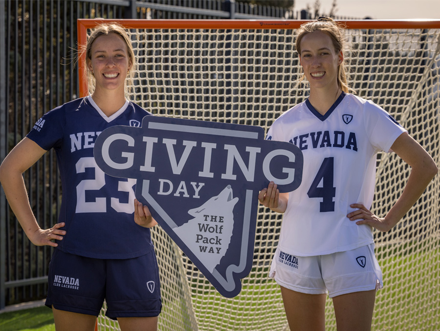 Two student athletes stand in front of a lacrosse goal holding a &ldquo;Giving Day&rdquo; sign and smiling.